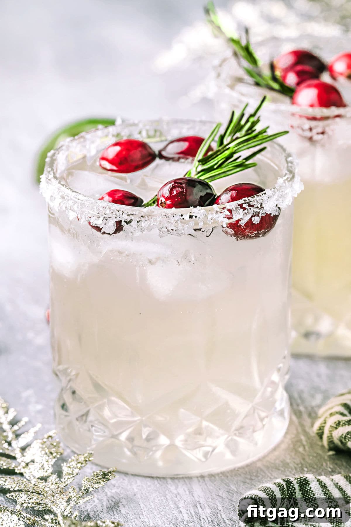 A frosty White Christmas Margarita in a short glass, beautifully topped with fresh red cranberries and a sprig of fragrant green rosemary. Another garnished glass is visible in the soft background, hinting at a joyful holiday gathering.