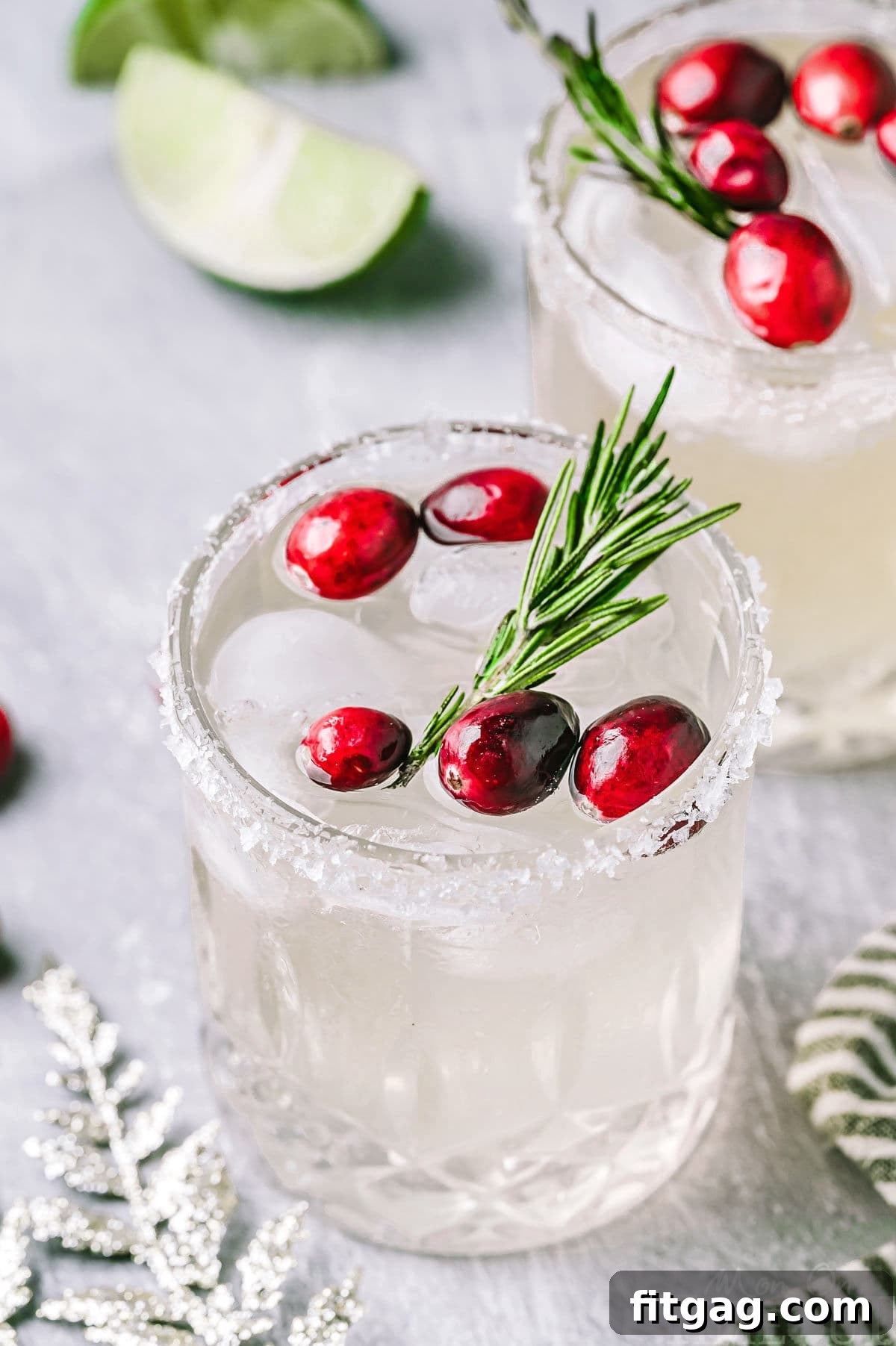 An overhead, angled view of two White Christmas Margaritas in elegant short glasses, each featuring a perfectly sugared rim. The drinks are garnished with glistening fresh cranberries and fragrant rosemary sprigs, ready for holiday enjoyment.