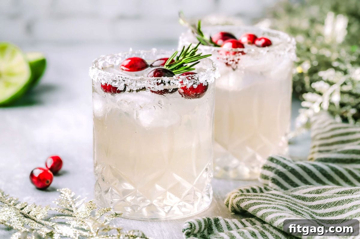 Two perfectly prepared White Christmas Margaritas, garnished with rosemary and cranberries, are placed beside a kitchen towel. Fresh lime wedges and additional cranberries are artfully scattered in the background, highlighting the ingredients.