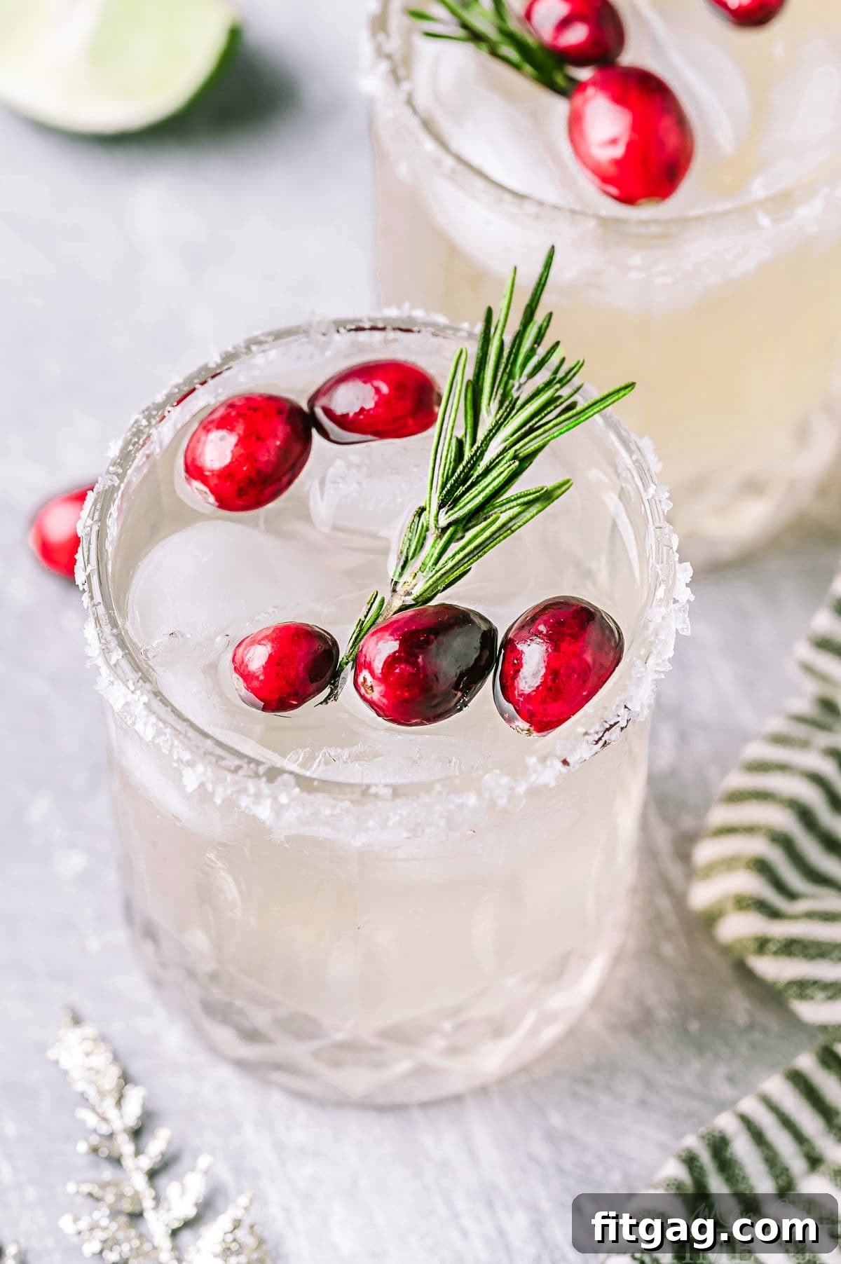 An angled down shot of two Christmas margaritas, served in short glasses with a sugared rim. The drinks are garnished with fresh cranberries and aromatic rosemary sprigs, creating a perfect festive tableau.