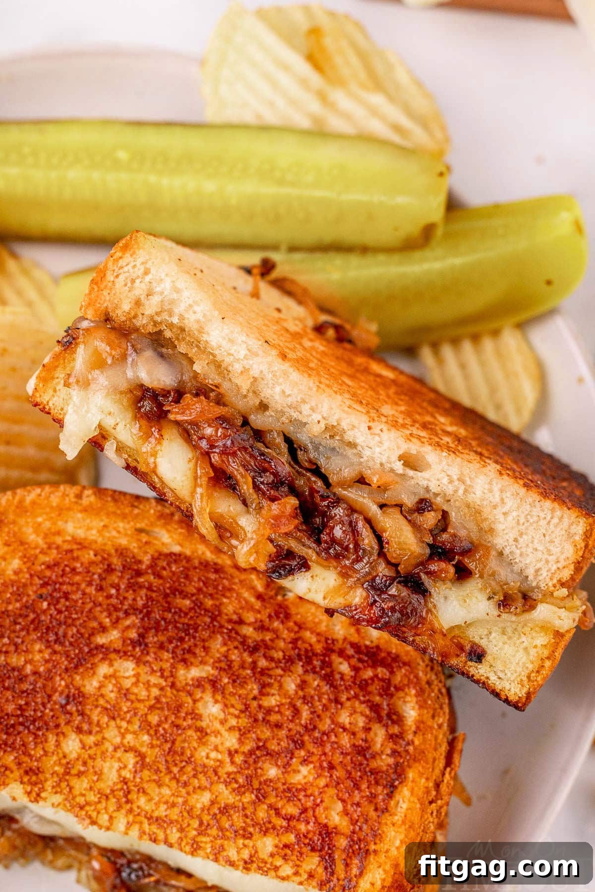 A beautifully presented French Onion Soup Grilled Cheese sandwich, cut in half, resting on a white plate alongside crisp pickle spears and a side of potato chips, ready to be enjoyed.