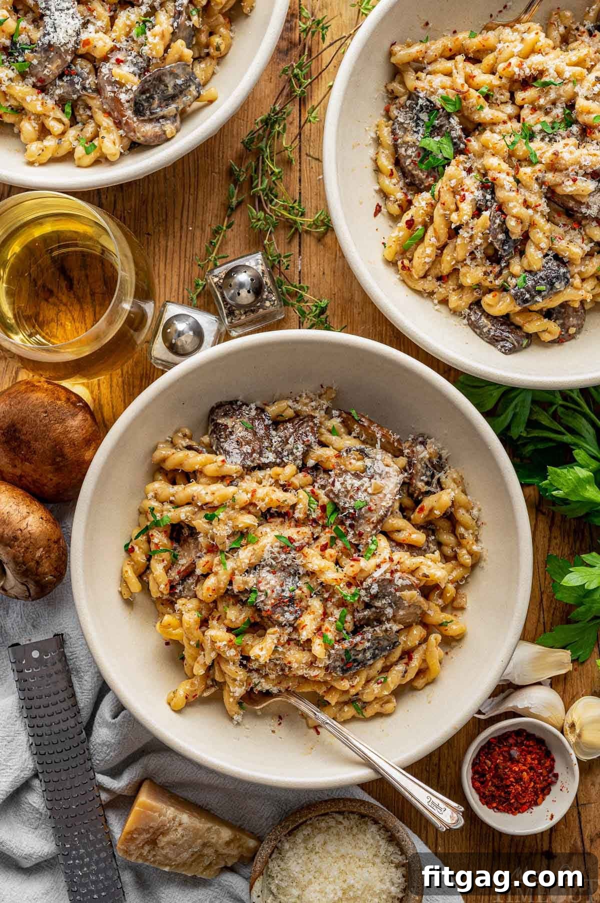 Top down shot of three bowls filled with a creamy mushroom pasta recipe. The bowls are arranged on a rustic wood surface, complemented by a glass of wine, fresh mushrooms, Parmesan cheese, and sprigs of herbs, creating an inviting and wholesome meal scene.