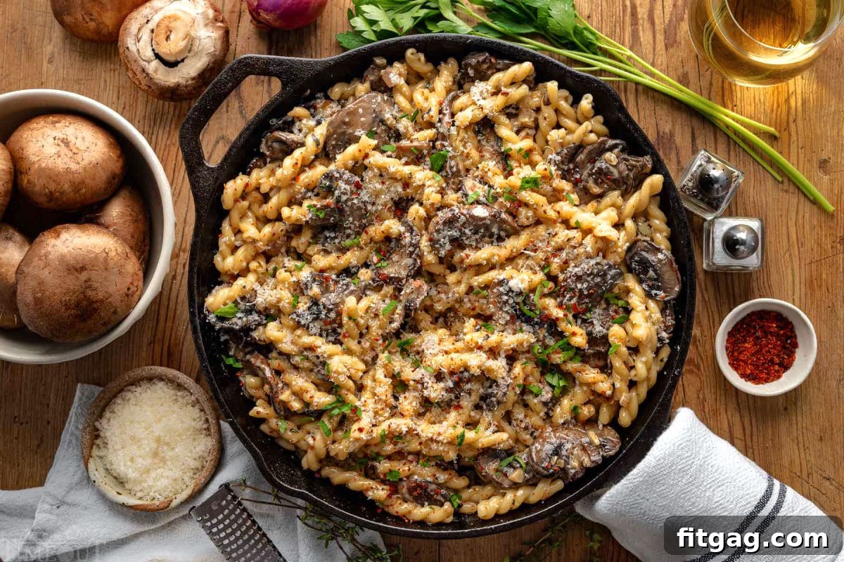 Top down view of a cast iron skillet filled with creamy mushroom pasta, generously garnished with fresh parsley. The skillet is artfully placed on a rustic wood surface, surrounded by perfectly arranged fresh mushrooms, shallots, and herbs, creating a harmonious and vibrant culinary display.
