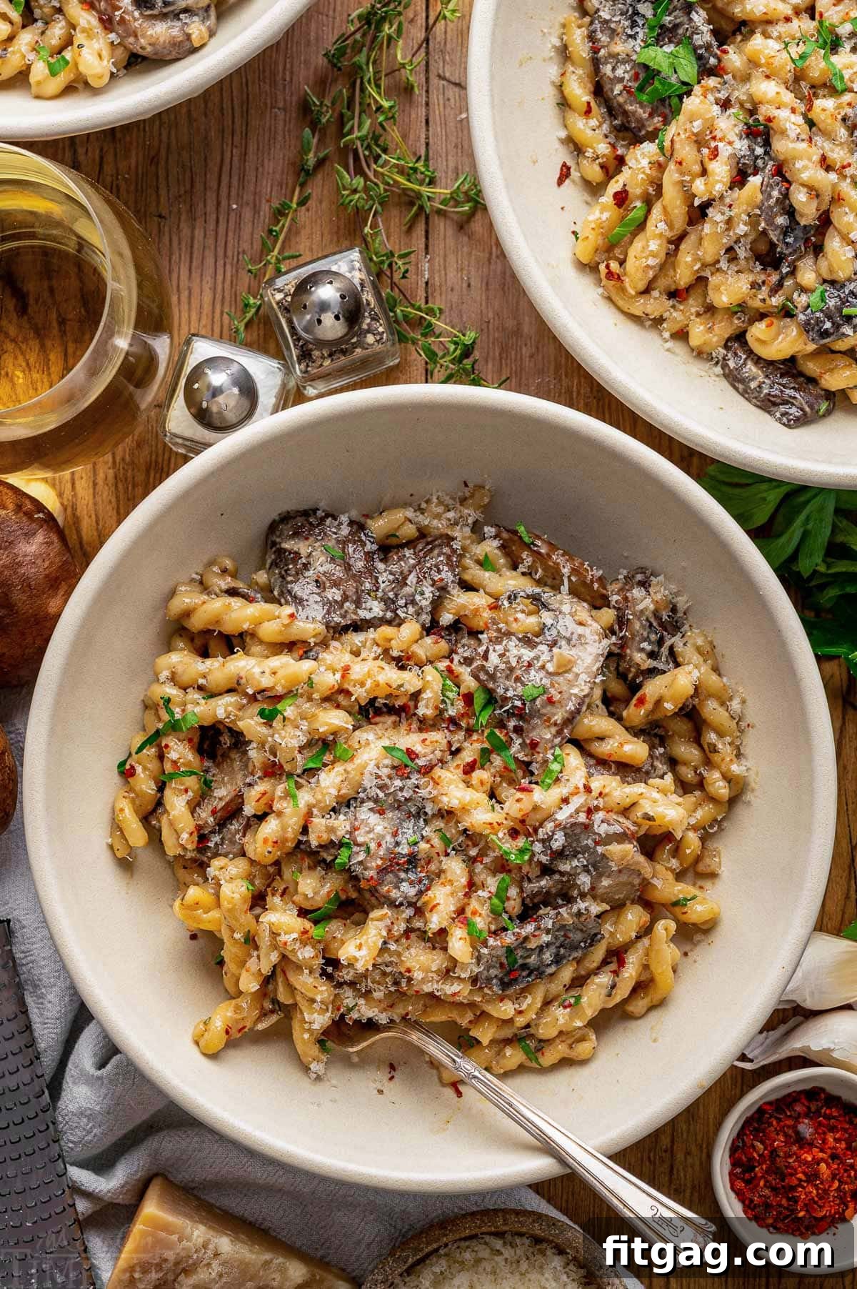 Top down shot of a white bowl filled with a creamy mushroom pasta recipe. The bowl is positioned on a wooden surface, accompanied by a glass of wine, two additional empty bowls, fresh Parmesan cheese, and sprigs of herbs, setting a charming and complete meal scene.