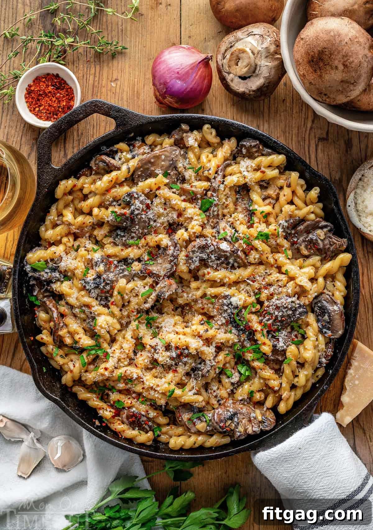 Top down view of a cast iron skillet filled with creamy mushroom pasta, finished with a fresh sprinkle of parsley. The skillet is invitingly placed on a wooden surface, accompanied by fresh mushrooms, shallots, and herbs, creating a mouthwatering display of ingredients.