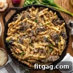 Top down view of cast iron skillet filled with mushroom pasta and finished with fresh parsley. Skillet is sitting on wood surface with mushrooms, shallots and herbs next to it.