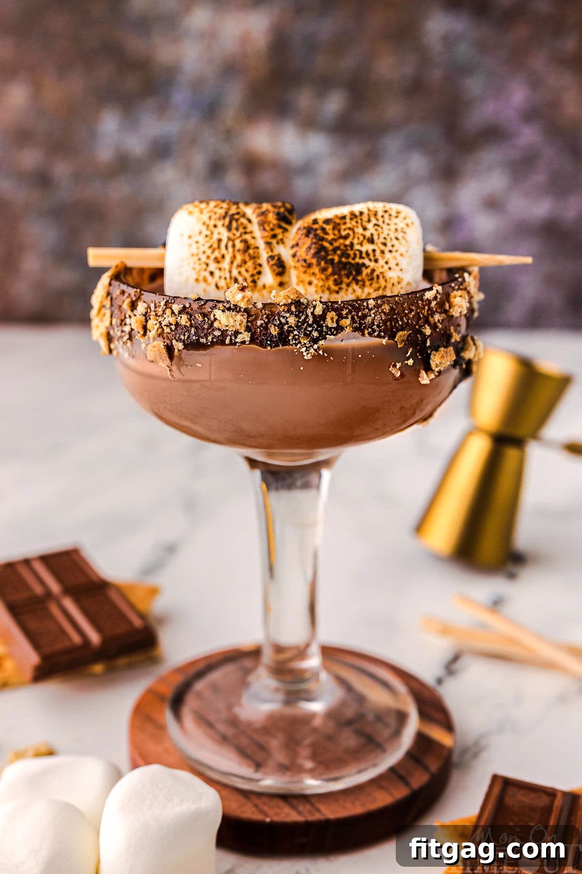 Side view of smores martini in a tall glass sitting on a wood coaster. Recipe ingredients are laying on the surface nearby.