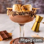 Side view of smores martini in a tall glass sitting on a wood coaster. Recipe ingredients are laying on the surface nearby.
