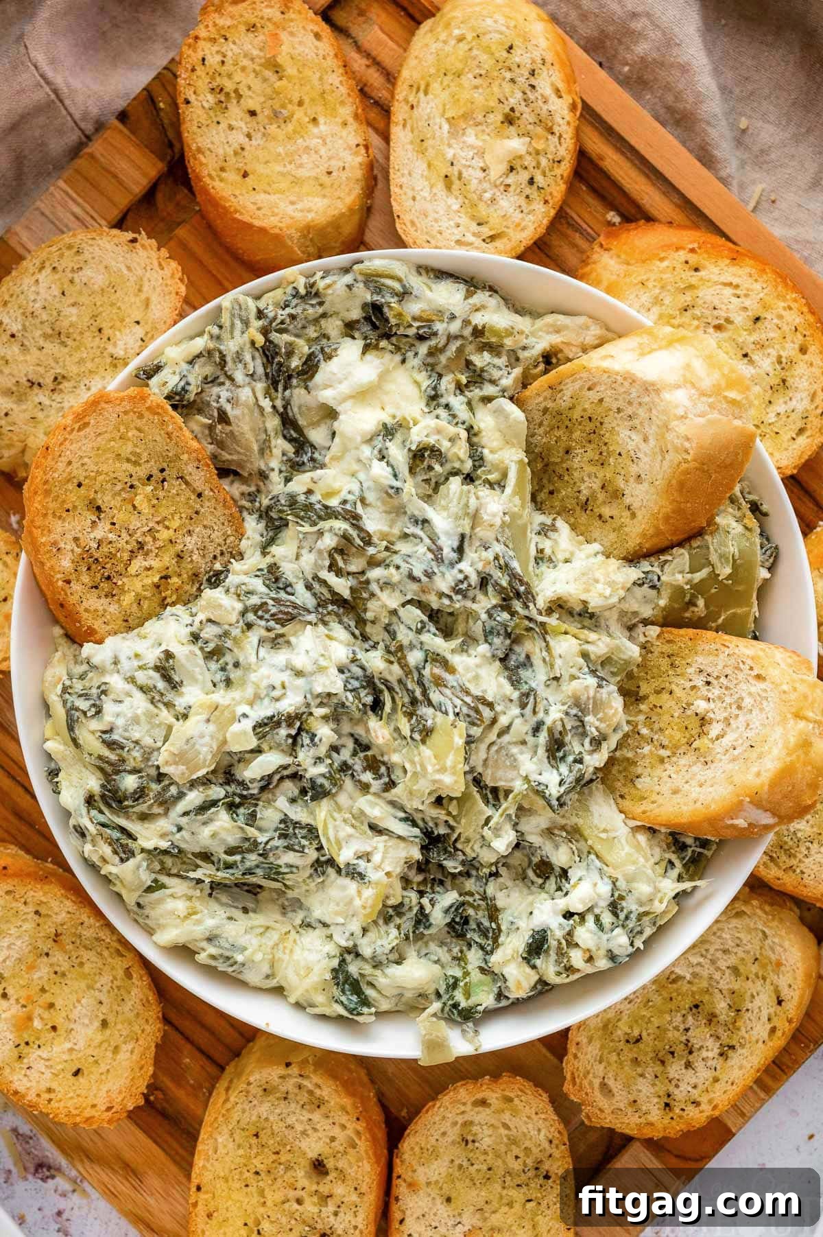 One Pot Wonder Spinach Artichoke Dip 2 Creamy Crockpot spinach artichoke dip in a white serving bowl, garnished and surrounded by golden brown crostini, ready for a party.