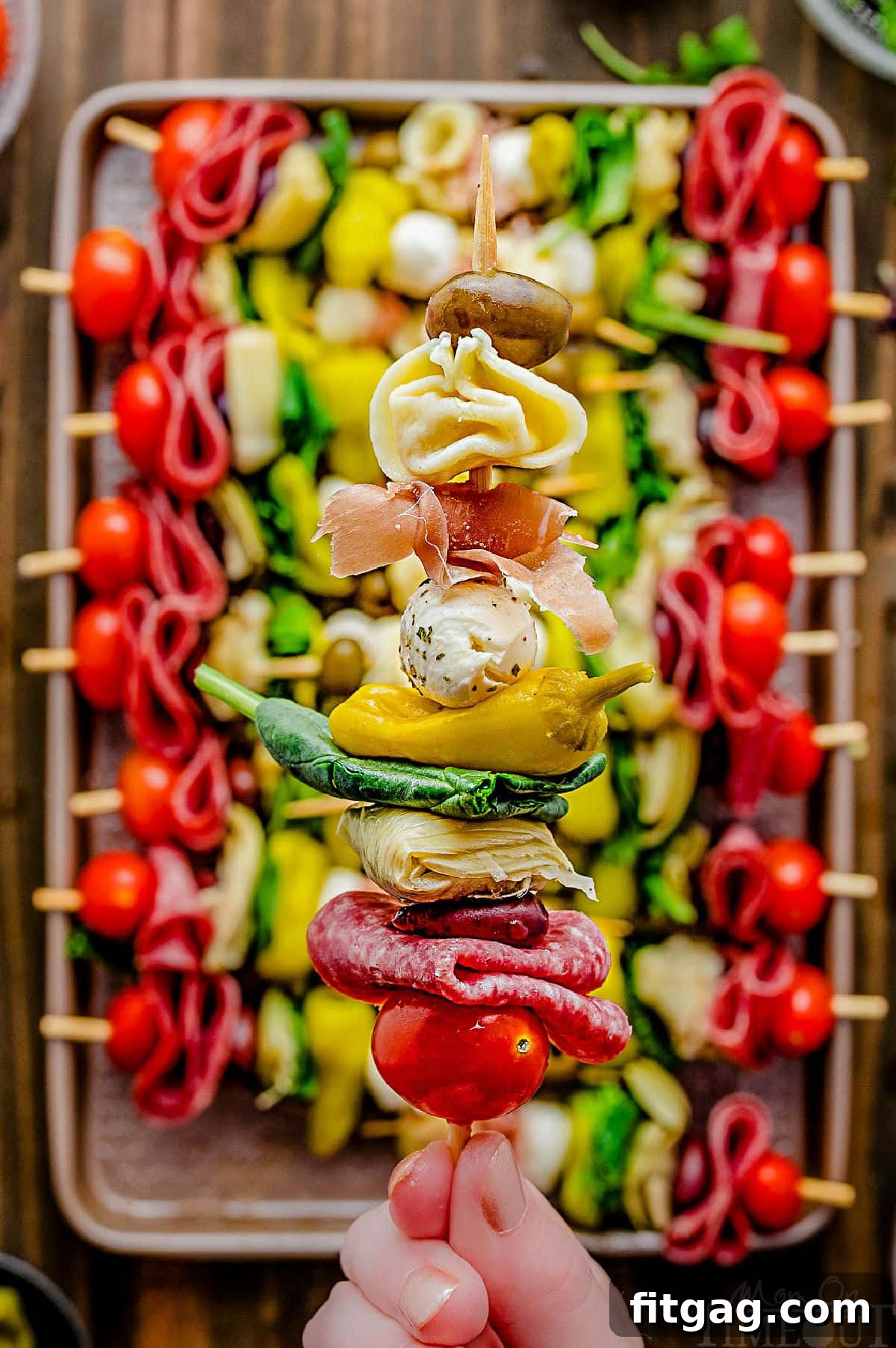 A single antipasto skewer held up, with a sheet tray filled with more colorful skewers in the background.