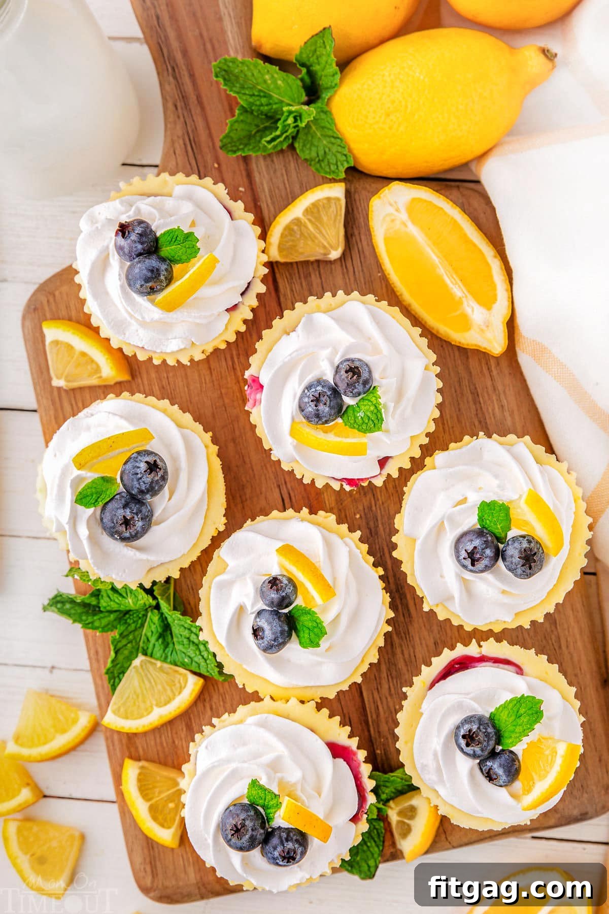 A top down view of lemon blueberry mini cheesecakes on a wood cutting board. The cutting board also has mint leaves and lemon slice garnishes on it.