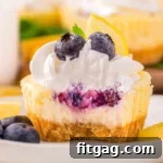 A close up view of a lemon blueberry mini cheesecake with a bite taken out of it. The cheesecake is on a white plate next to garnishes.