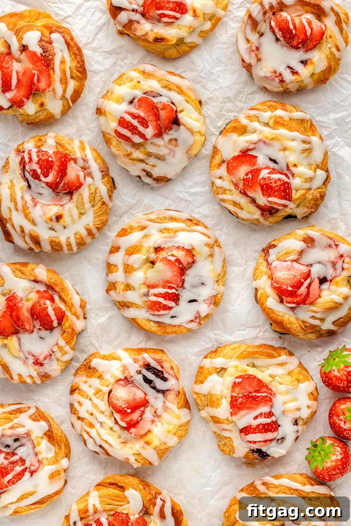 Strawberry Danish Delight 3 An overhead view of numerous Strawberry Cream Cheese Danishes arranged neatly on parchment paper, showcasing their round, twisted pastry edges and generous strawberry and cream cheese fillings.