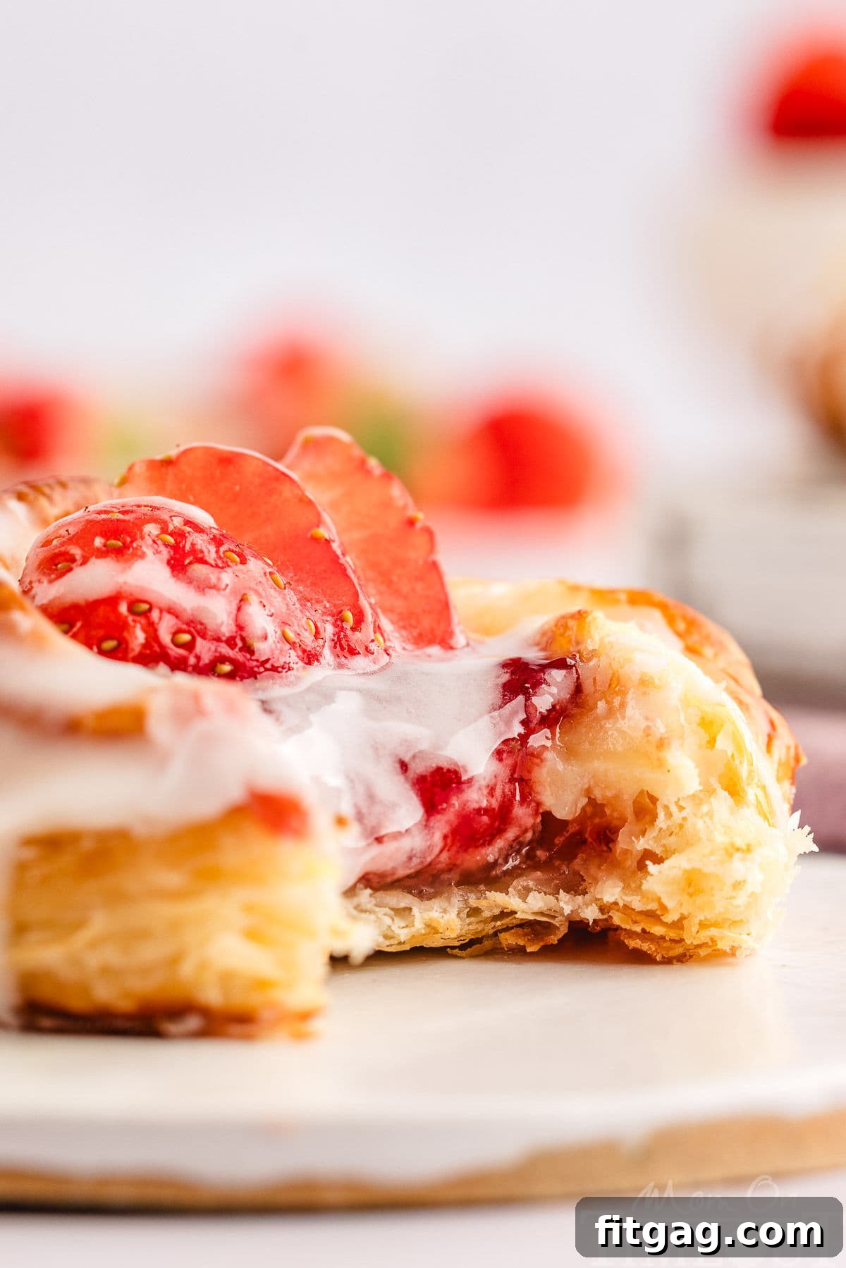 Strawberry Danish Delight 7 A close-up of a Strawberry Cream Cheese Danish on a white wooden board, showing a bite taken out, revealing the flaky pastry and creamy filling, with glaze and fresh strawberries on top.
