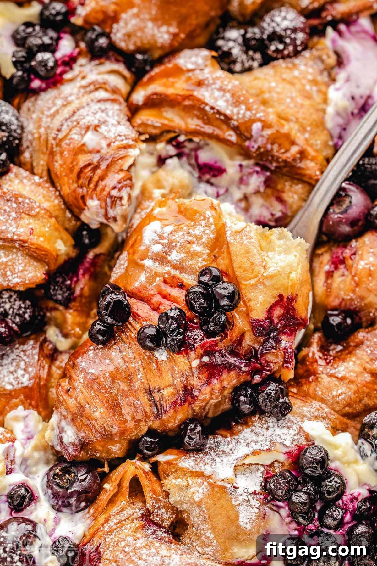 A piece of golden-brown blueberry croissant French toast bake being lifted out of a casserole dish with a serving spatula.