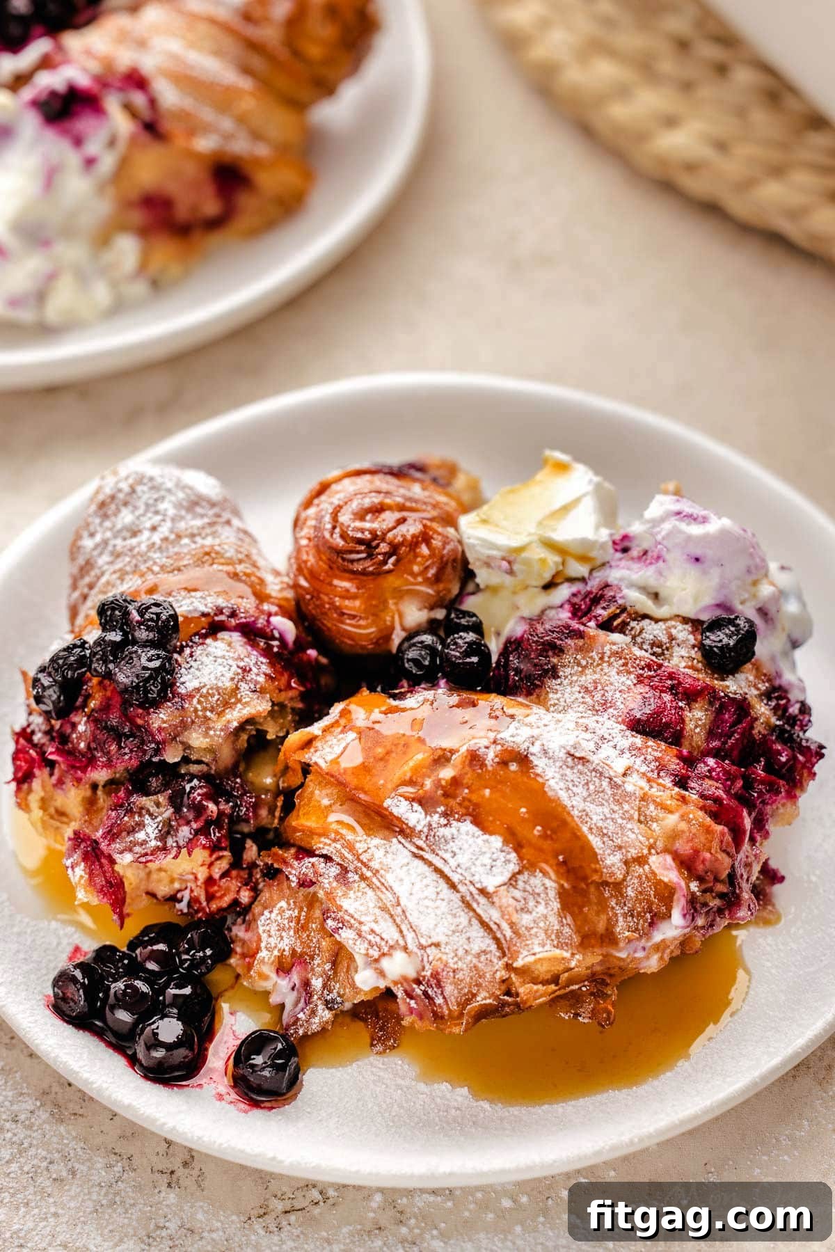 A serving of Blueberry Croissant French Toast Bake on a white round plate, topped with a generous drizzle of maple syrup and fresh blueberries.