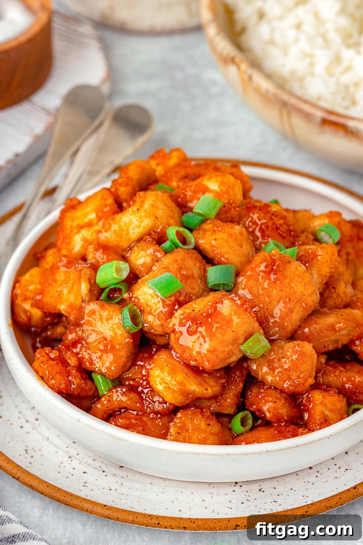 A pristine white bowl brimming with delectable Firecracker Chicken, garnished with fresh green onions. In the soft-focus background, a companion bowl of fluffy white rice awaits.