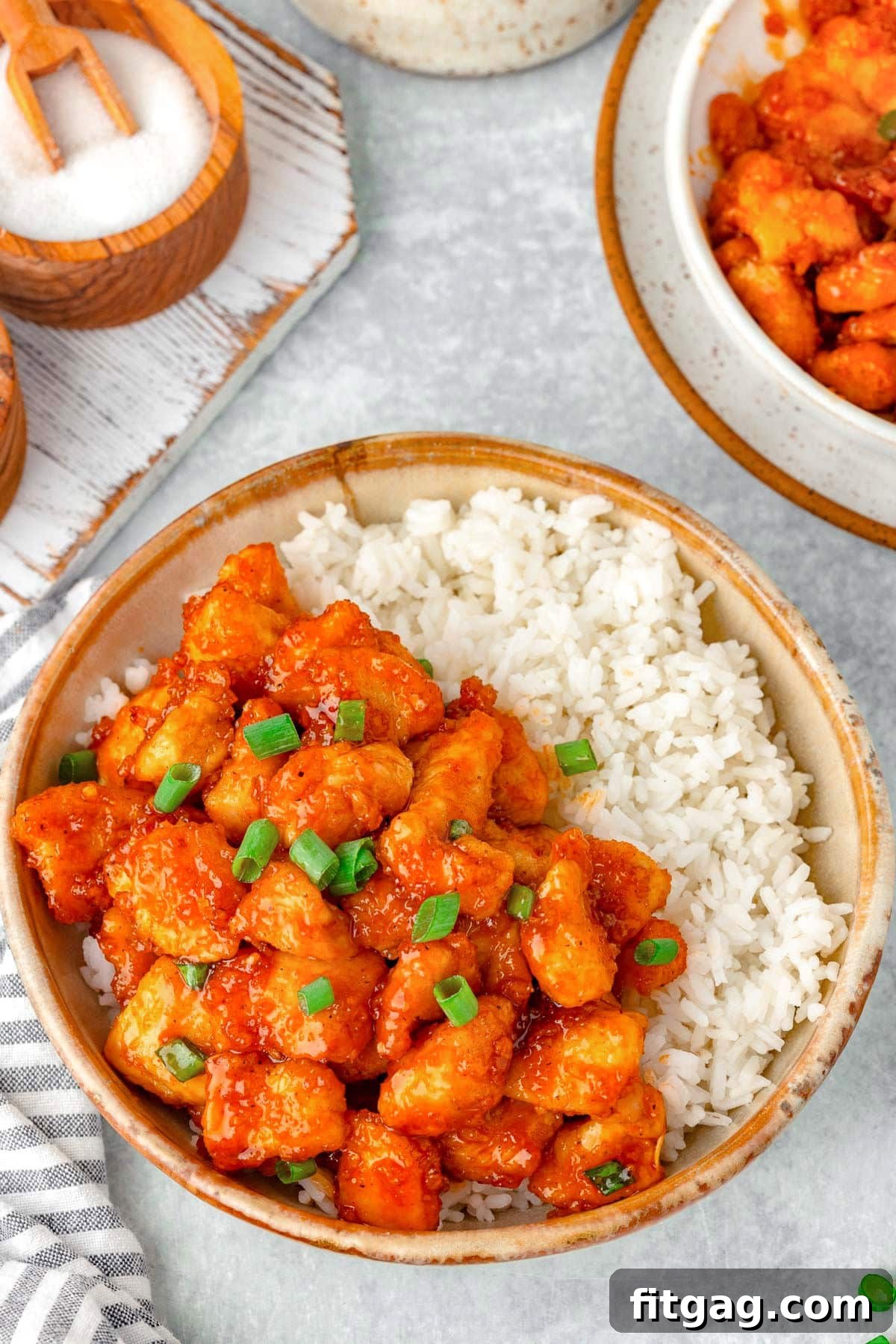 A top-down perspective capturing a bowl of pristine white rice alongside a generous serving of Firecracker Chicken. A subtly patterned towel rests nearby, completing the inviting setup.
