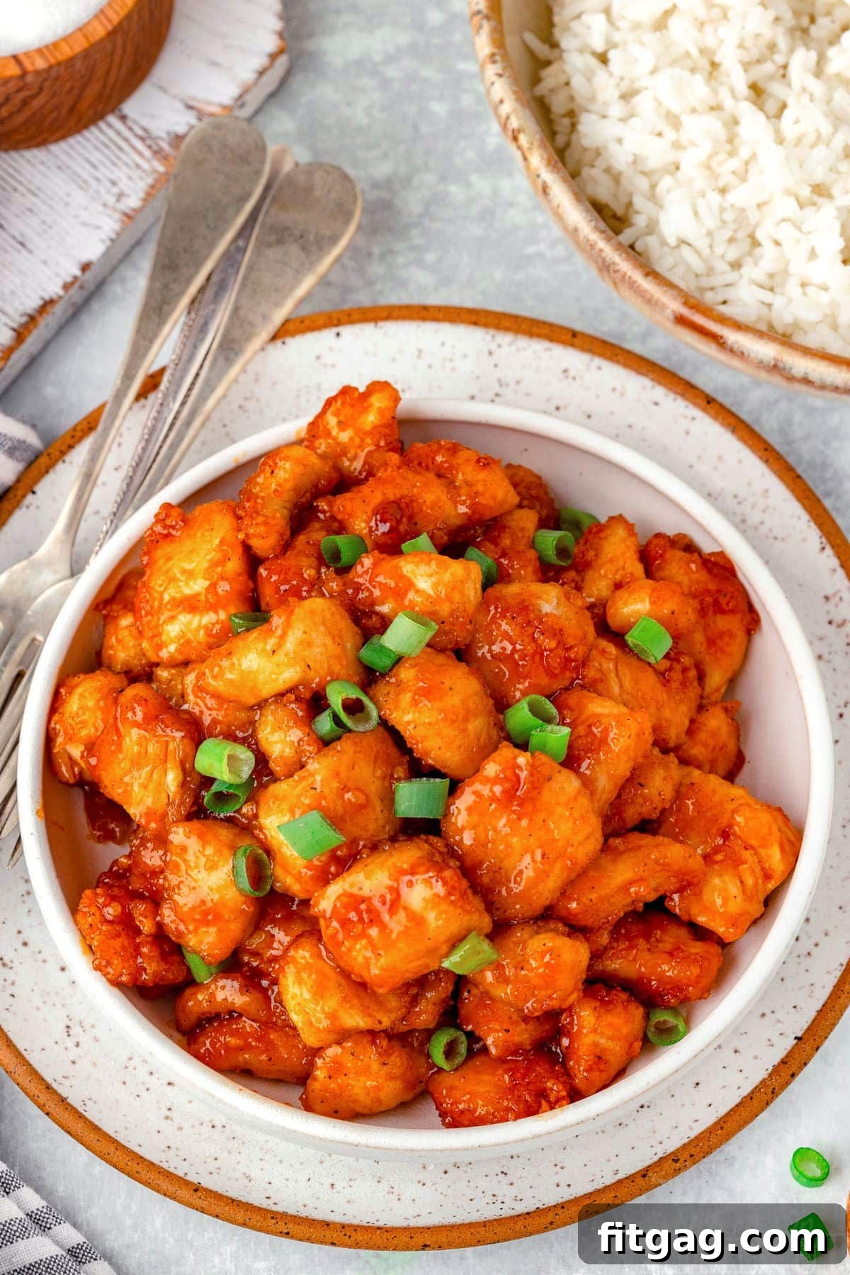 A stunning shot of a white bowl overflowing with Firecracker Chicken, perfectly garnished with sliced green onions, with a bowl of fluffy rice gently blurred in the background, highlighting the main dish.