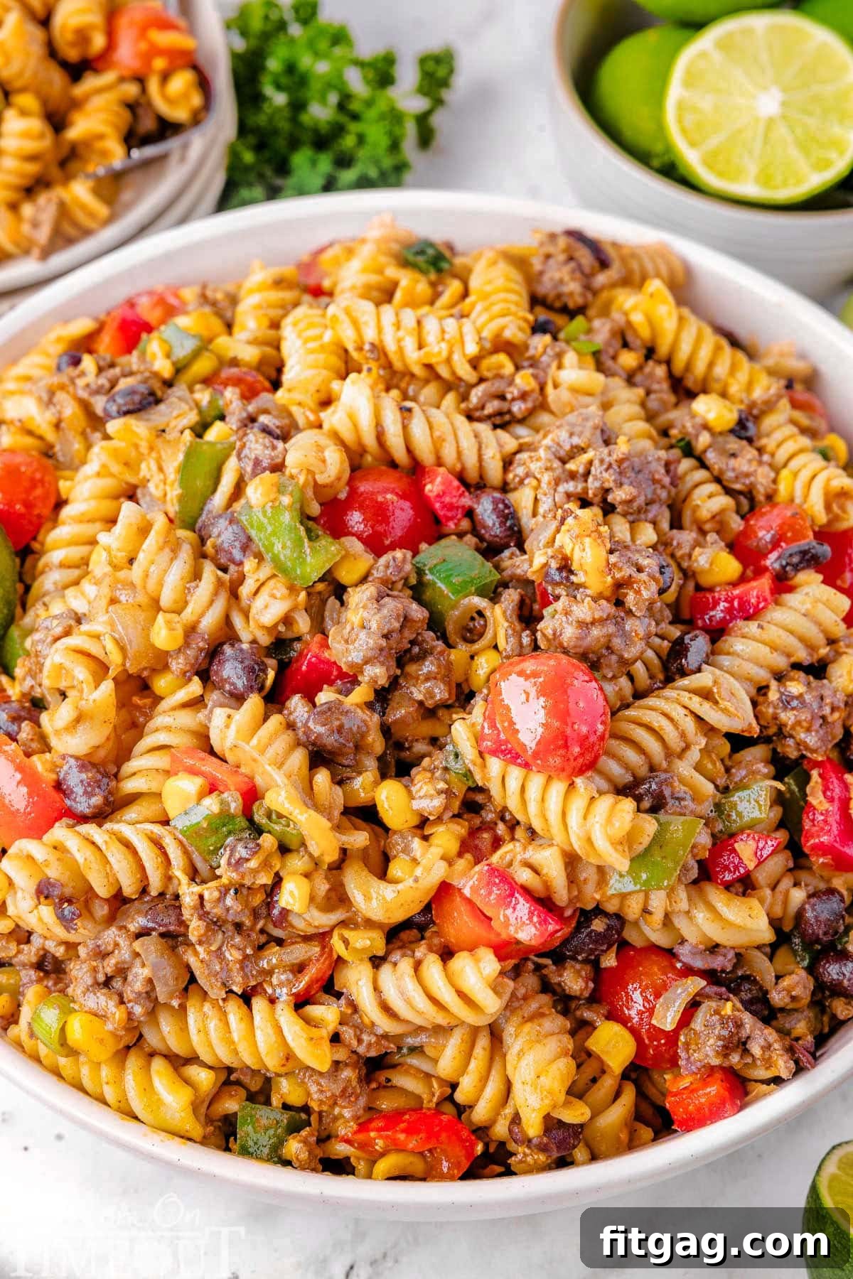 Chuckwagon Pasta Salad 2 A large serving bowl overflowing with vibrant Cowboy Pasta Salad, garnished with fresh lime wedges. In the background, a plate of the salad is ready to be enjoyed, alongside a few whole limes, suggesting its zesty flavor.