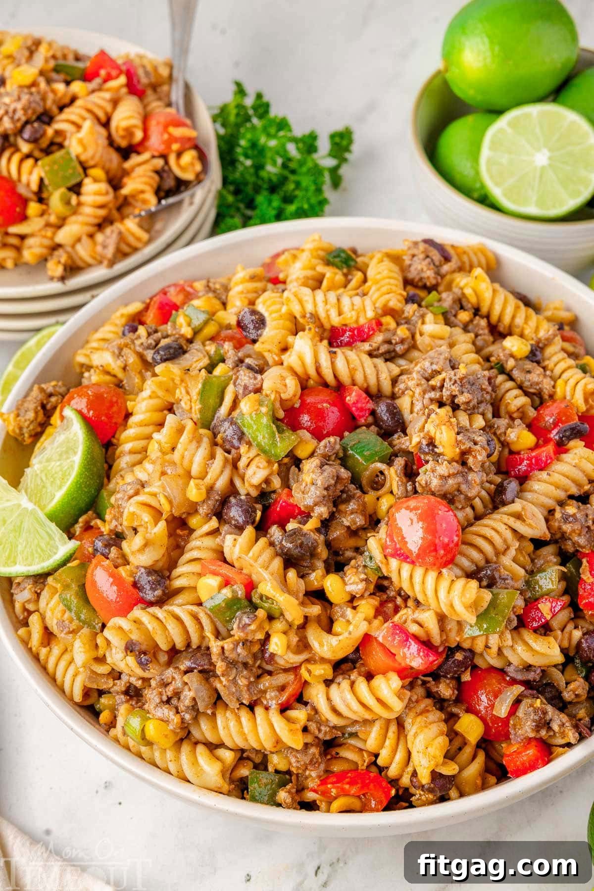 Chuckwagon Pasta Salad 8 Cowboy pasta salad in a white serving bowl garnished with lime wedges. A stack of plates topped with salad is visible in the background, ready for guests.