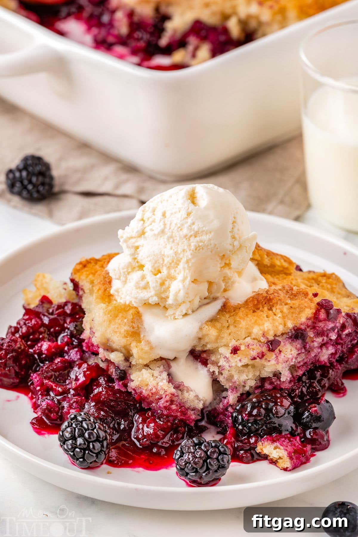 Sweet Berry Indulgence 2 Berry cobbler served on white round plate and topped with vanilla ice cream. Baking dish with the rest of the berry cobbler recipe can be seen in the background.