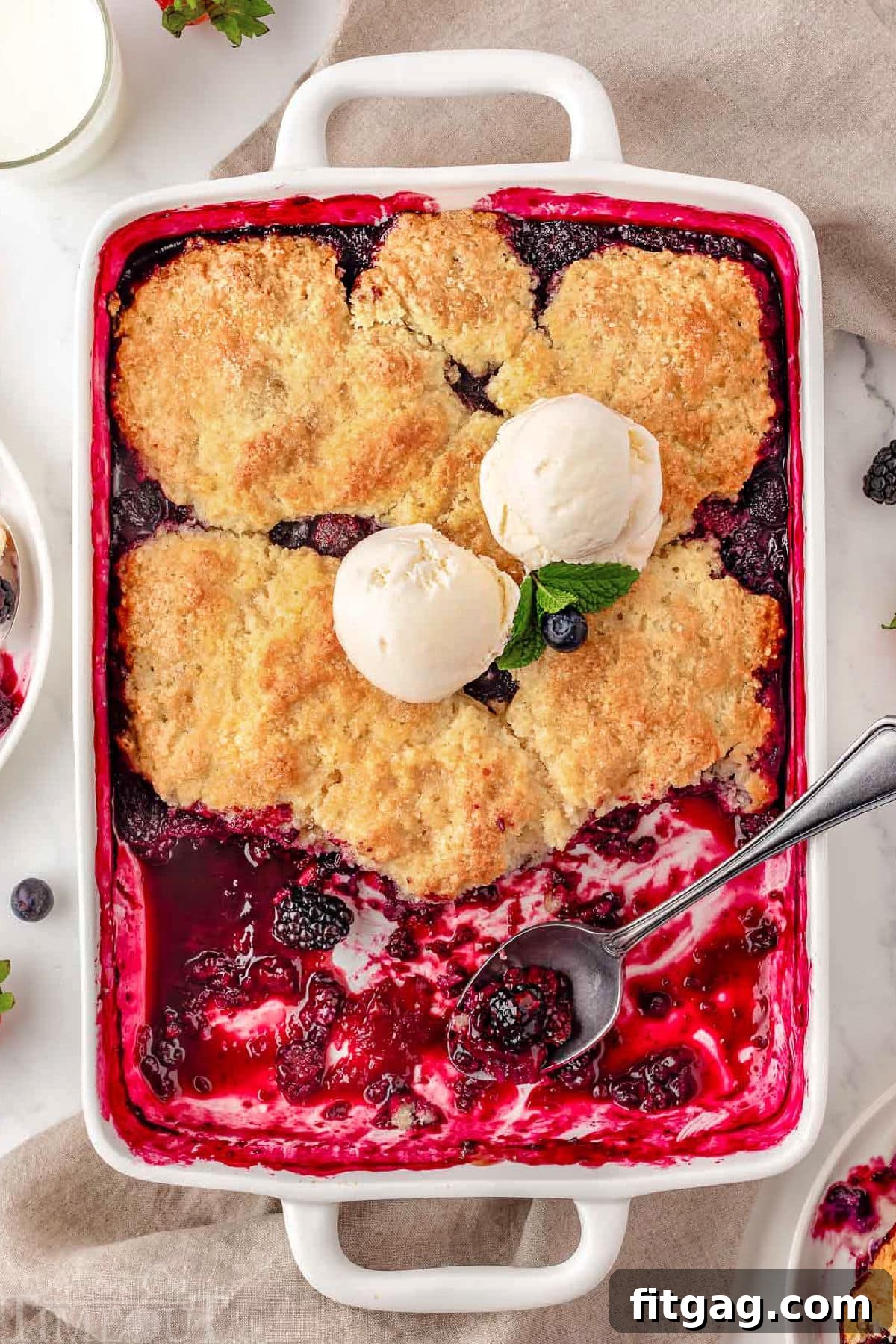 Sweet Berry Indulgence 9 Top down shot of berry cobbler recipe in white baking dish with some servings of the cobbler removed. Topped with vanilla ice cream and fresh mint leaves.