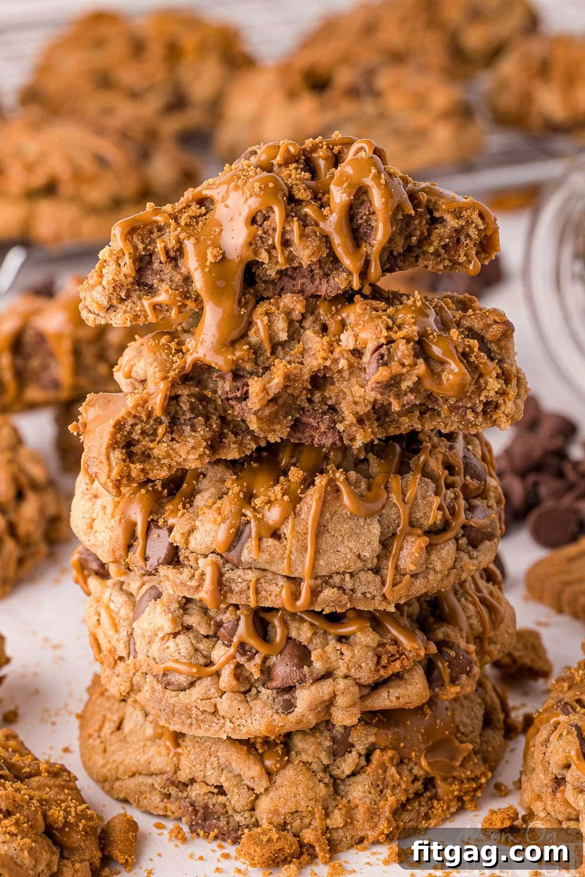 Golden Speculoos Chocolate Chip Dreams 8 A stack of four Biscoff chocolate chip cookies, with the top one broken in half to showcase its texture. They are drizzled with cookie butter and placed on white parchment, with more cookies in the background.