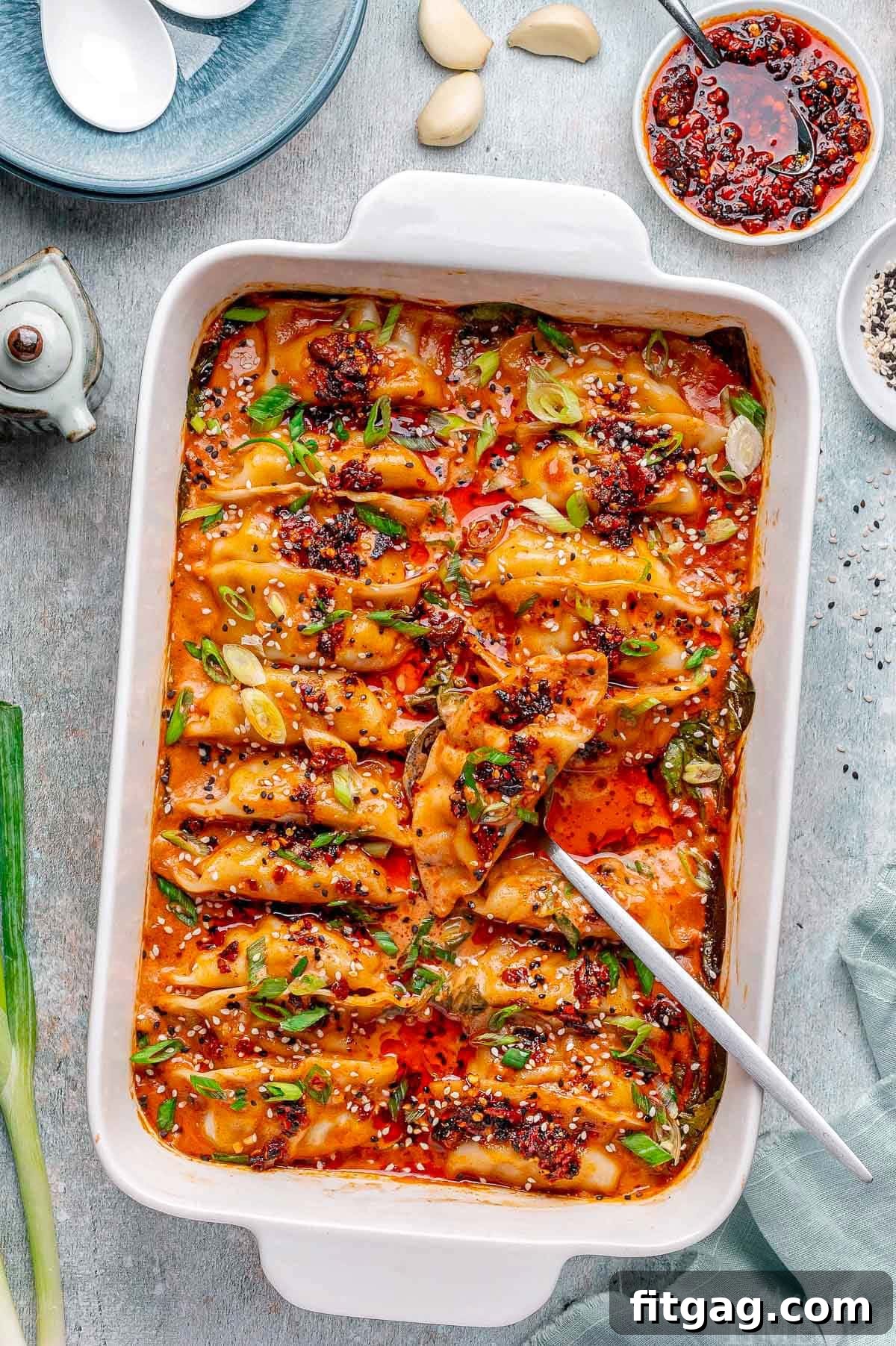 Overhead shot of vibrant baked dumplings in a white baking dish, elegantly garnished with chili crisp, fresh green onions, and a sprinkle of black and white sesame seeds. A spoon rests in the dish, inviting diners to serve themselves.
