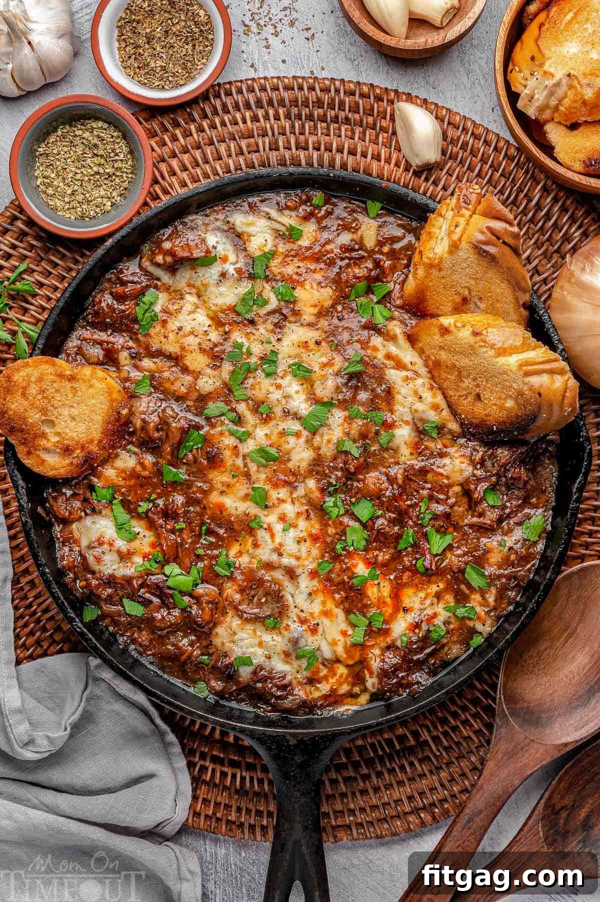 Slow-Cooked French Onion Beef 2 Overhead shot of french onion pot roast in cast iron skillet with cheese on top and baguette slices scattered about. Fresh parsley garnishes the dish.