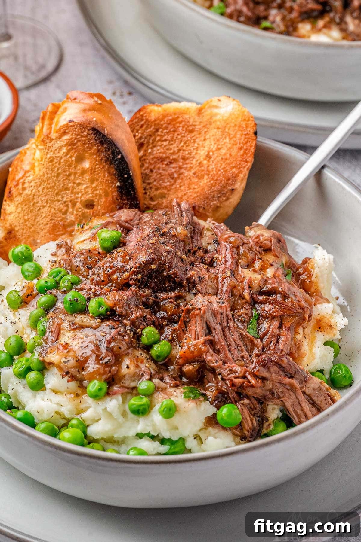 Slow-Cooked French Onion Beef 7 Close up shot of french onion soup pot roast in bowl with mashed potatoes, peas and french bread.
