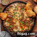Overhead shot of french onion pot roast in cast iron skillet with cheese on top and baguette slices scattered about. Fresh parsley garnishes the dish.
