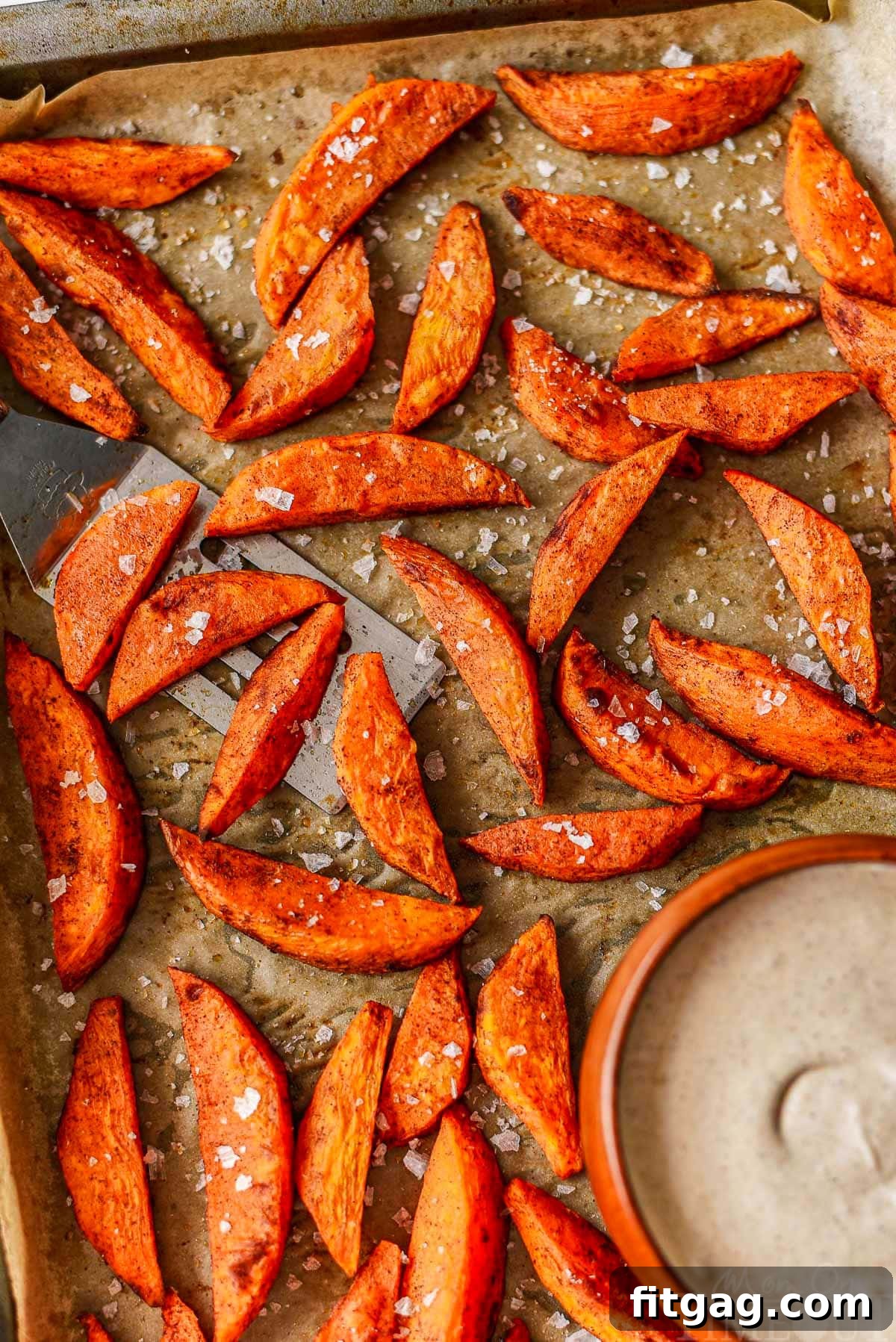 Top-down view of roasted sweet potato wedges on a parchment-lined baking sheet, alongside a bowl of rich brown sugar cinnamon marshmallow dip.