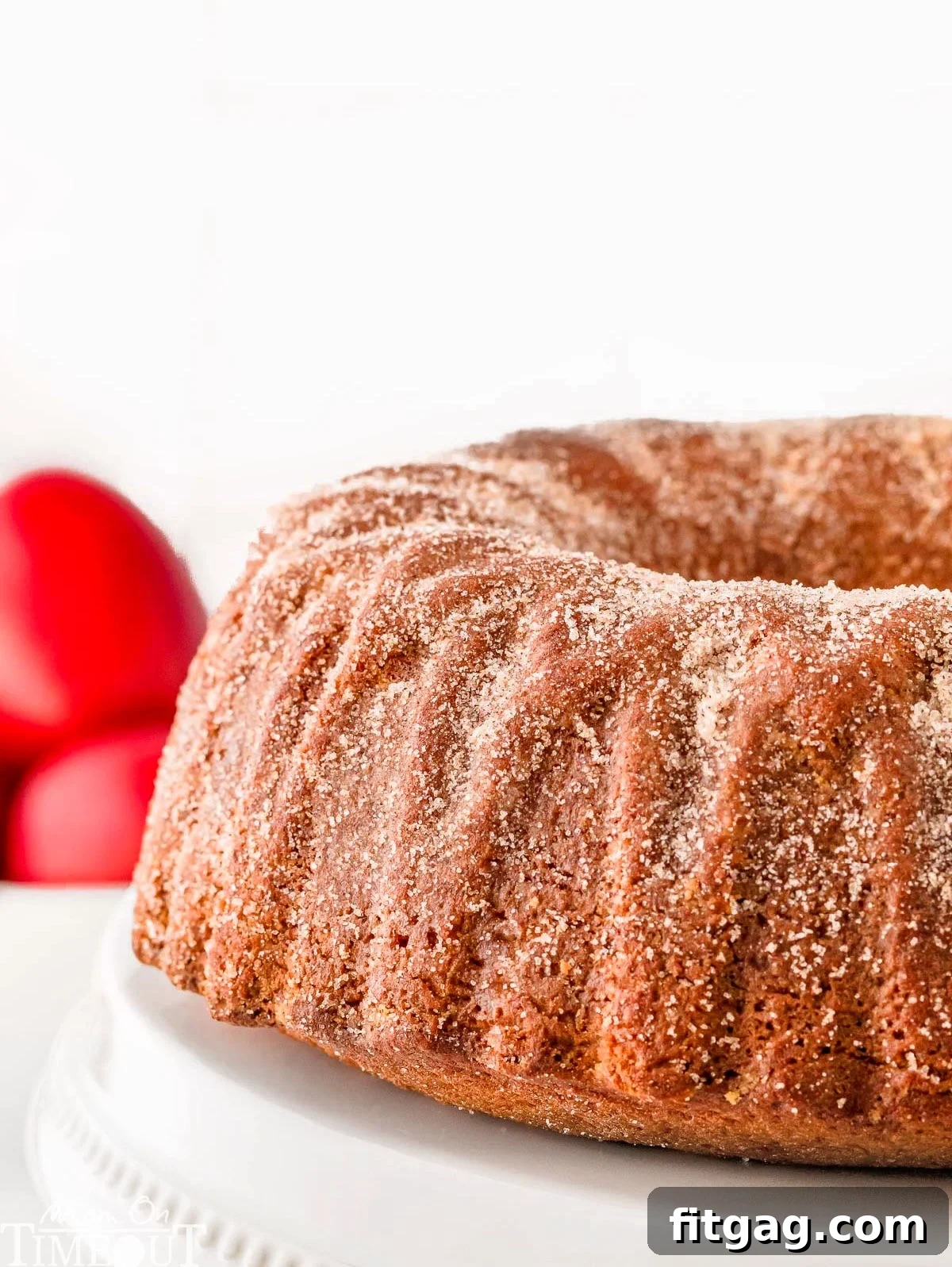 A stunning side view of a whole, golden-brown apple cider bundt cake, presented on an elegant white cake stand, with vibrant red apples artfully arranged in the background, evoking a perfect autumn scene.