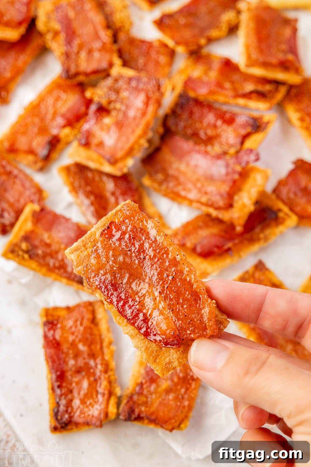 A single candied bacon cracker held above a pile of more crispy crackers on white parchment paper, ready to be enjoyed.