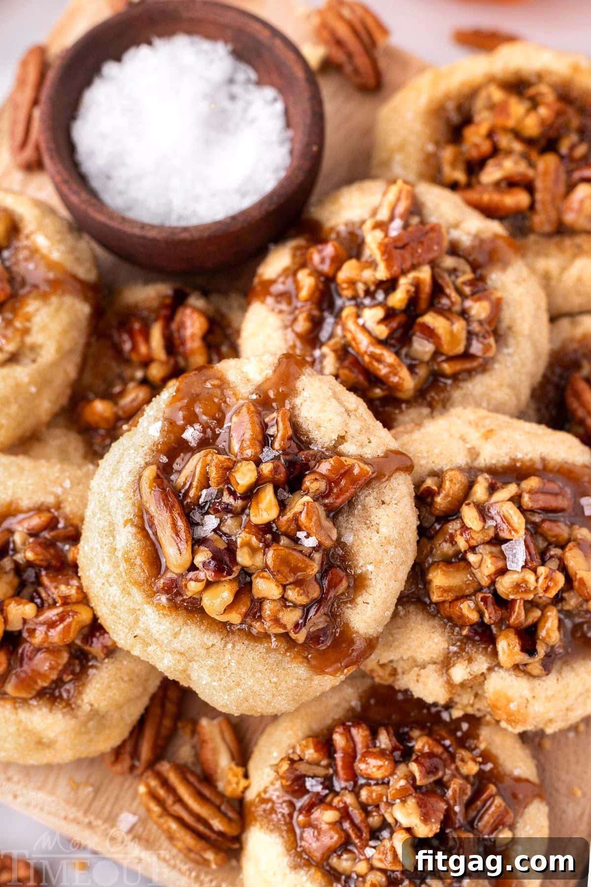 Overhead shot of pecan pie cookies piled high on a wood board next to a small bowl of flaky sea salt. 