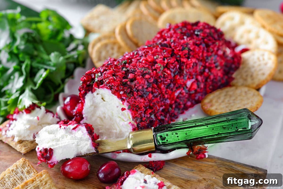 Wide shot of cranberry salsa coated goat cheese log on board with crackers. A green cheese knife is set beside the cheese log.