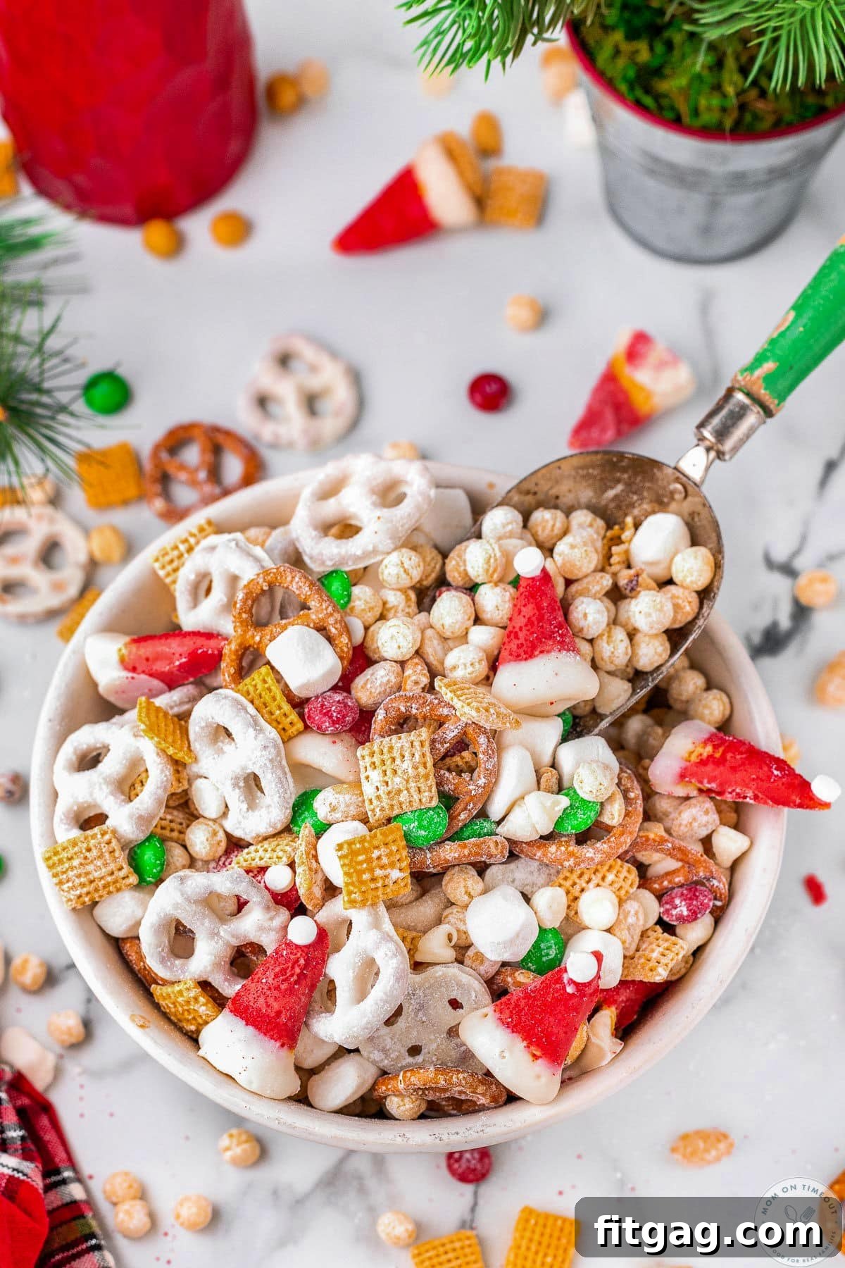 White bowl filled with christmas snack mix with small scoop. Pretzels, peanuts and candy are scattered about the bowl.