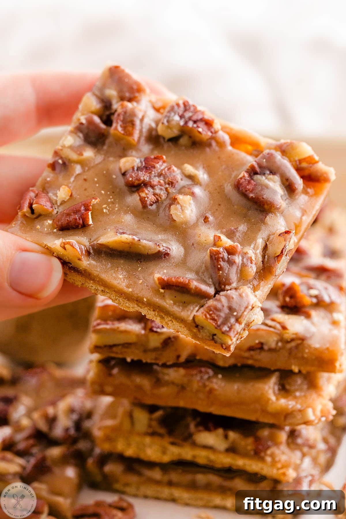 Toasted Pecan Brittle 8 A hand reaching to pick up a piece of pecan pie bark from a tempting stack, with scattered pecans in the background.