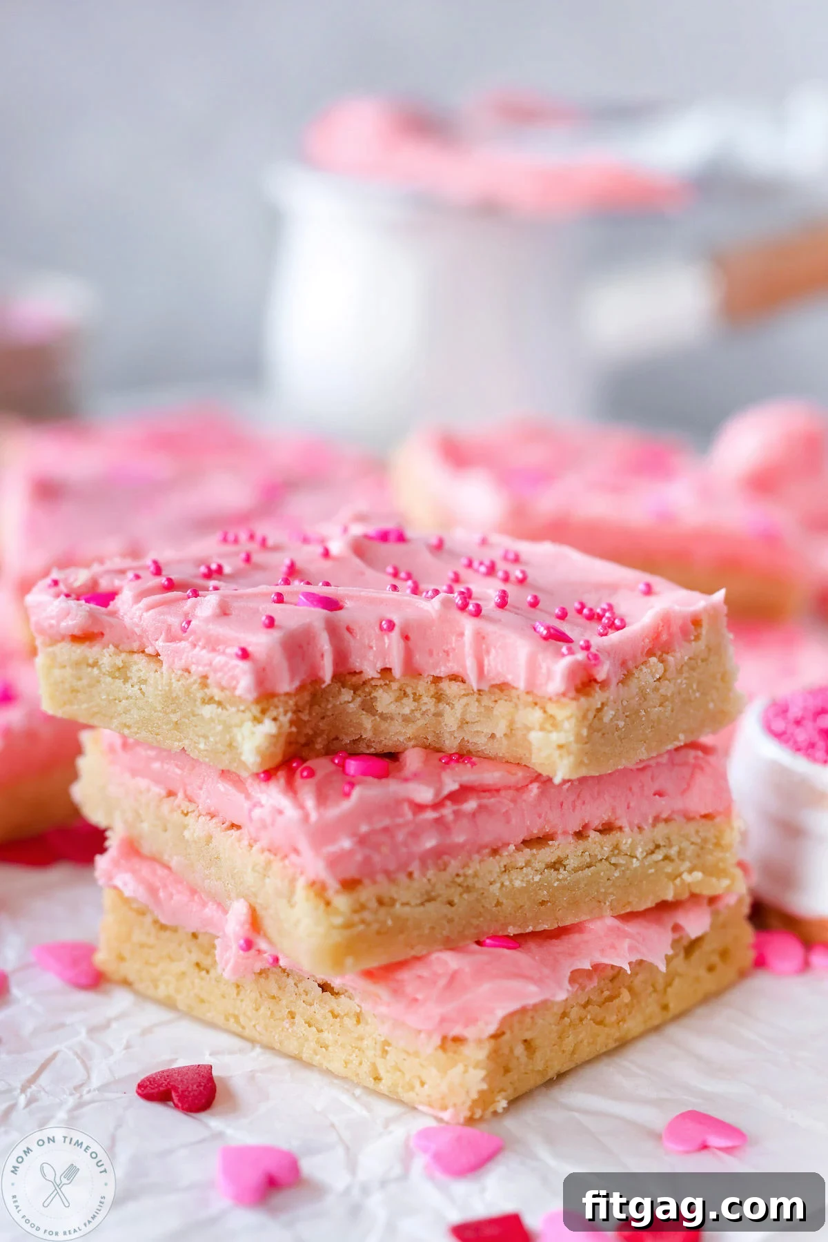 Sweetheart Sugar Cookie Bars 2 Three Valentine's Day sugar cookie bars frosted with pink cherry frosting and festive sprinkles, stacked on white parchment paper. The top bar has a bite taken out of it, revealing its soft interior.