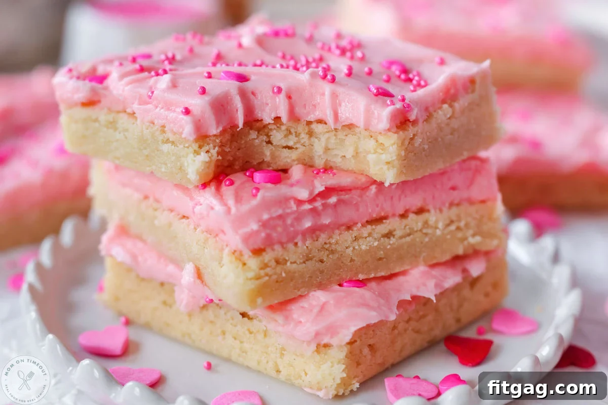 Sweetheart Sugar Cookie Bars 4 A close-up shot of three Valentine's Day sugar cookie bars, frosted with pink cherry frosting and adorned with heart-shaped sprinkles, stacked on a charming white scalloped-edge plate. The top bar shows a tempting bite taken out.