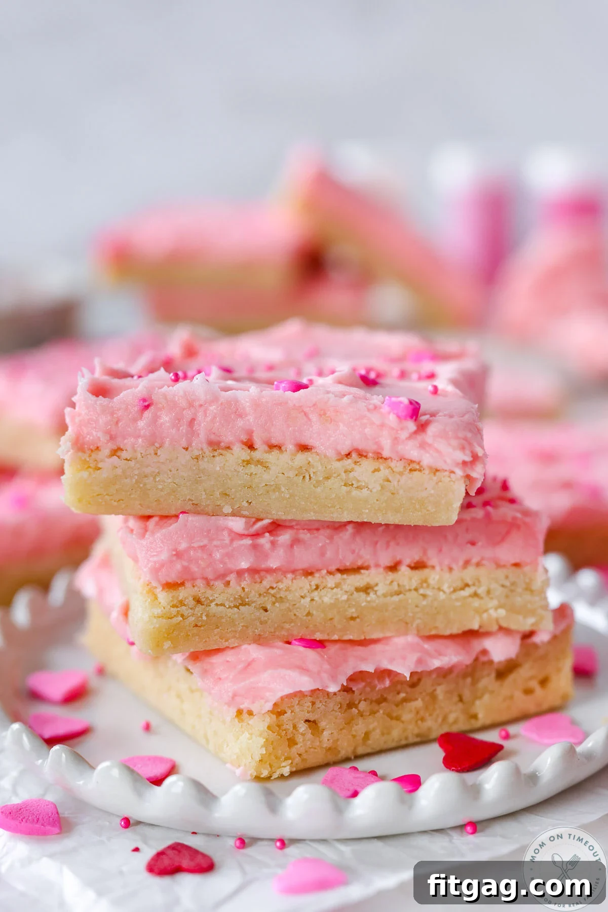 Sweetheart Sugar Cookie Bars 8 A stack of three delightful sugar cookie bars, beautifully decorated for Valentine's Day with pink frosting and festive sprinkles, resting on a round white plate.
