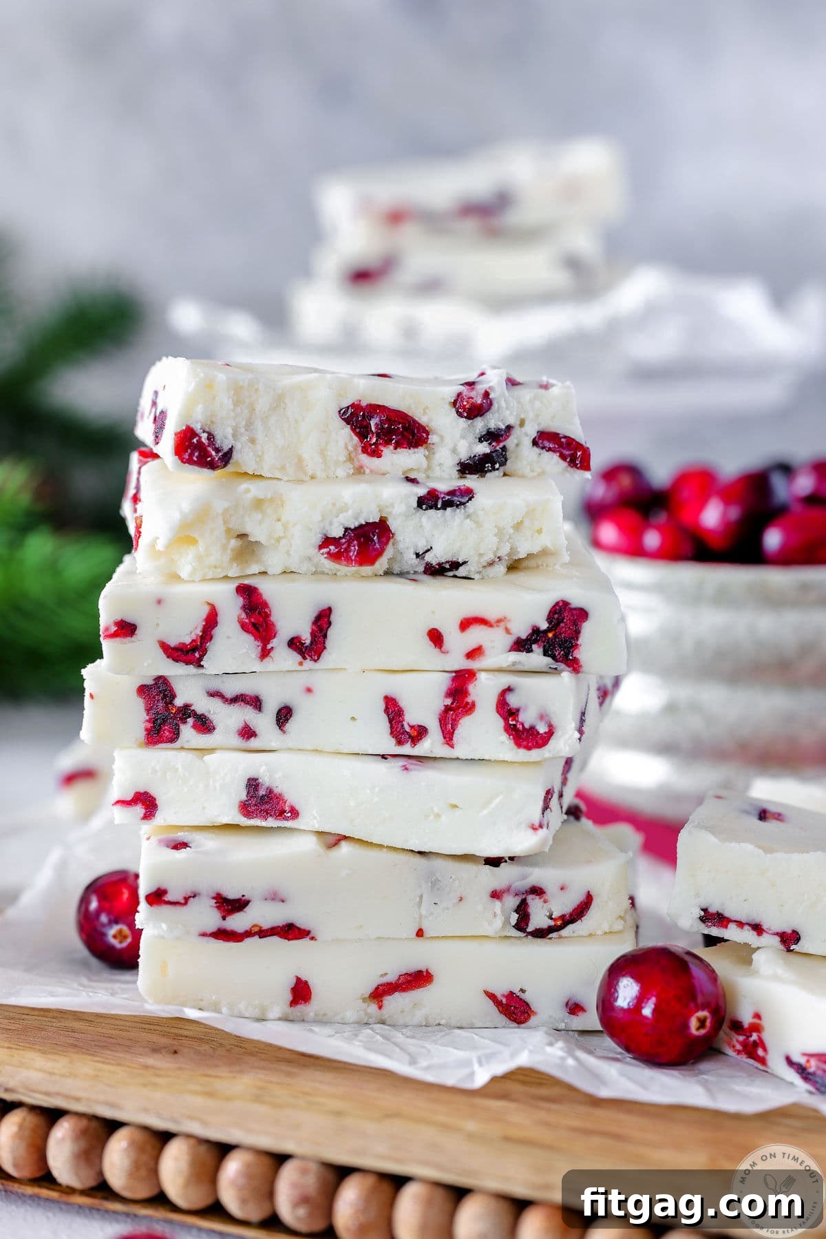 Tall stack of cranberry fudge on wood and marble cutting board. Fresh cranberries and more fudge can be seen in the background.
