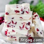 White chocolate cranberry fudge stacked high on vintage white pedestal. Greenery and fresh cranberries can be seen in background.