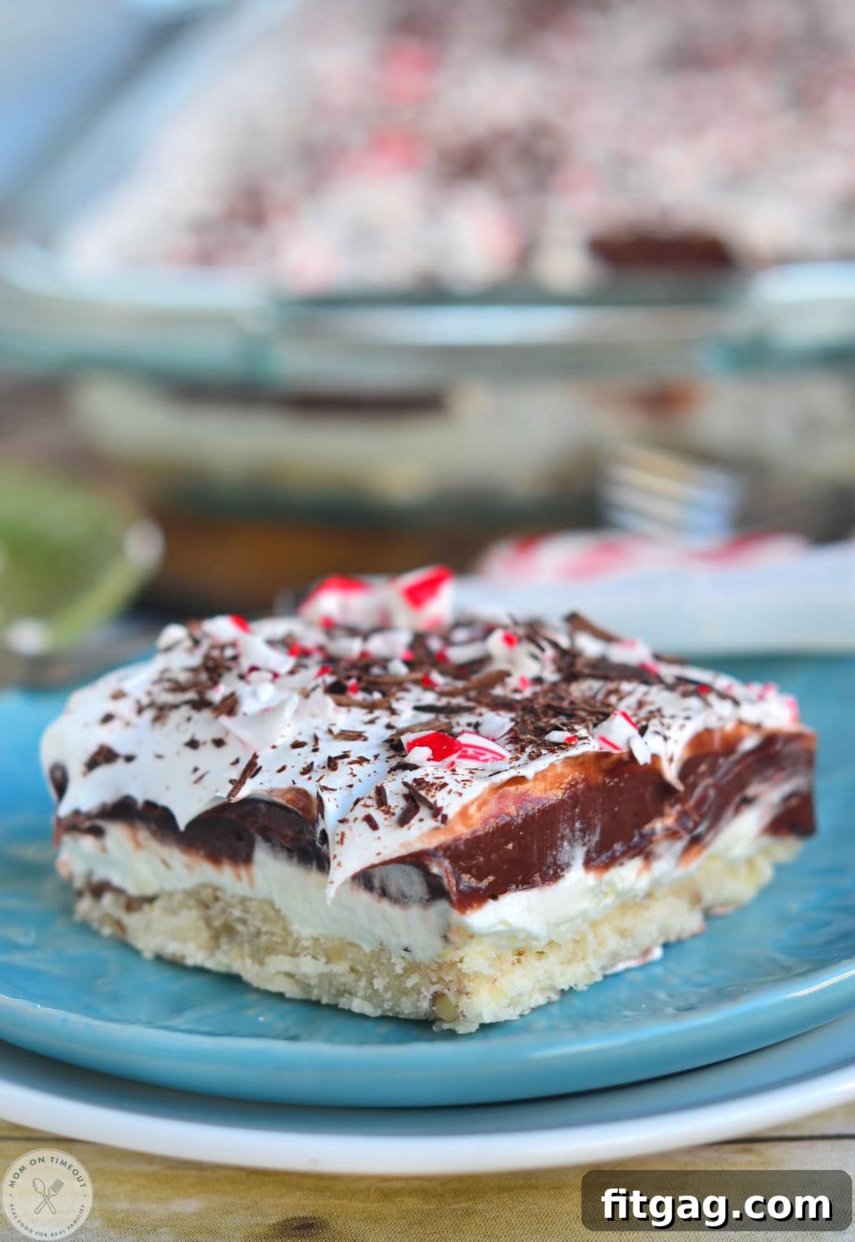A slice of four-layered chocolate peppermint dessert on a blue plate, garnished with crushed candy canes. The Peppermint Lush is topped with crushed candy canes.