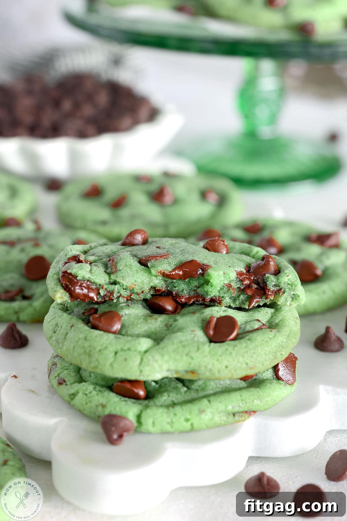 Two mint chocolate chip cookies with one half cookie on top with melty chocolate. Cookies are green with chocolate chips. Green cake stand can be seen in the background.