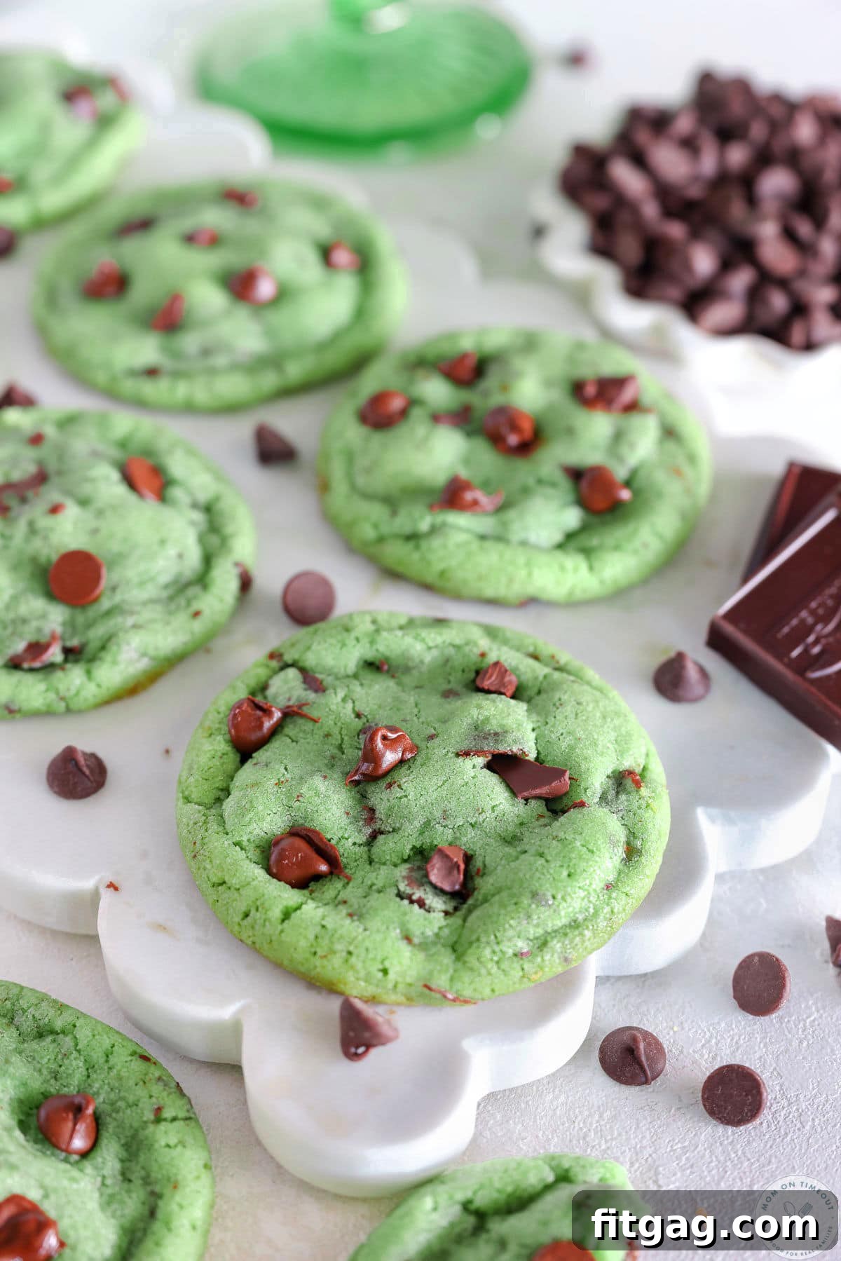 Four mint chocolate chip cookies baked and resting on white marble board with bowl of chocolate chips next to it. 