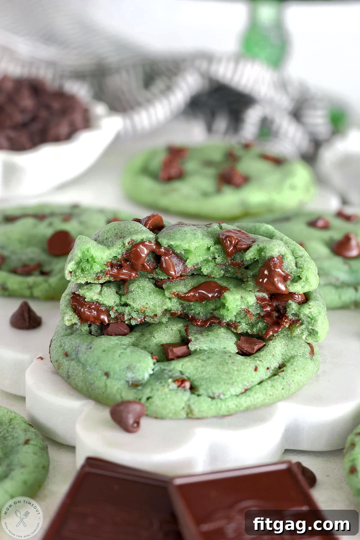 Stack of mint chocolate chip cookies on scalloped white marble board. Top cookie is cut in half.