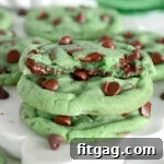 Two mint chocolate chip cookies with one half cookie on top with melty chocolate. Cookies are green with chocolate chips. Green cake stand can be seen in the background.