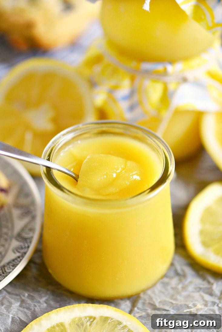 A close-up view of vibrant lemon curd inside a glass jar, with a spoon resting in it, surrounded by fresh lemon wedges, ready to be sampled.